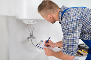 Male Plumber Writing On Clipboard In Front Of Electric Boiler In Kitchen Room